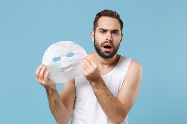 Bearded young man 20s years old in white shirt hold tissue fabric face mask isolated on blue pastel background, studio portrait. Skin care healthcare cosmetic procedures concept. Mock up copy space.