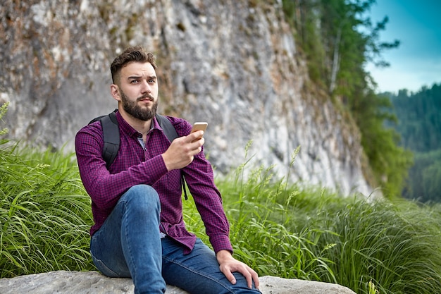 Bearded young man of caucasian ethnos holds smartphone in his hands, during adventures on hiking trail