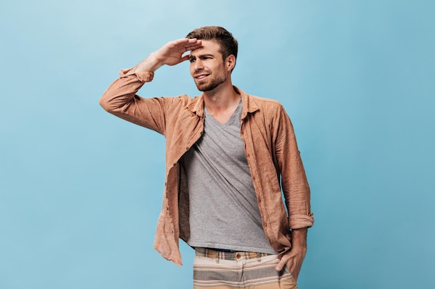 Bearded young man in grey cool tshirt beige shirt and striped modern trousers looking away on isolated blue background