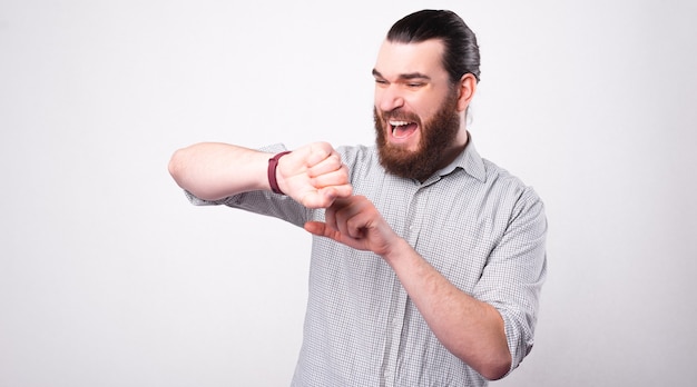 A bearded young man is looking stressed at his watch being afraid that he is late near a white wall