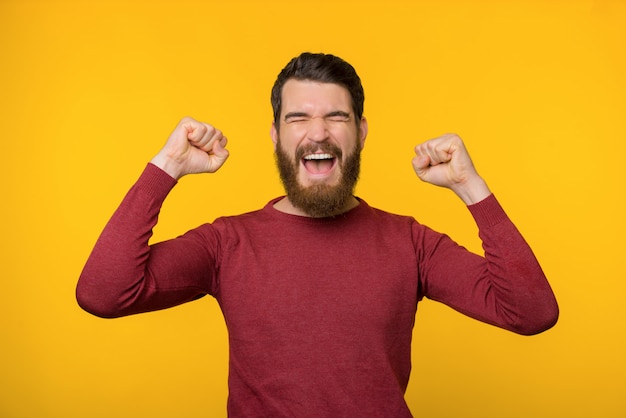 Bearded young man is screaming of joy with both of his hands up near yellow background.