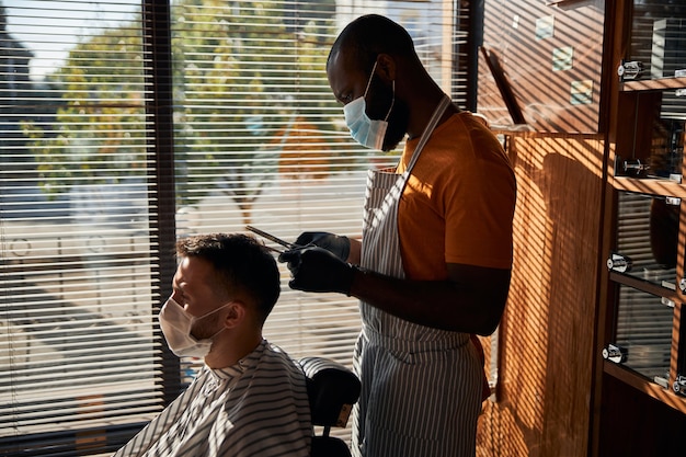 Bearded young man in medical mask getting haircut in barbershop