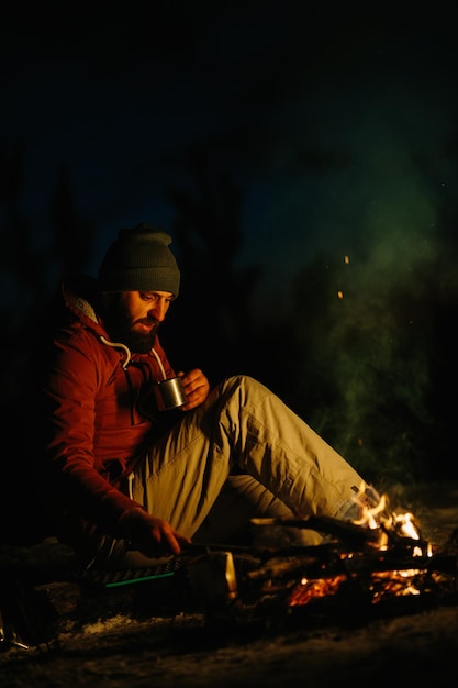 A bearded young man rests by the night campfire in the winter forest The concept of survival and hiking in the wild