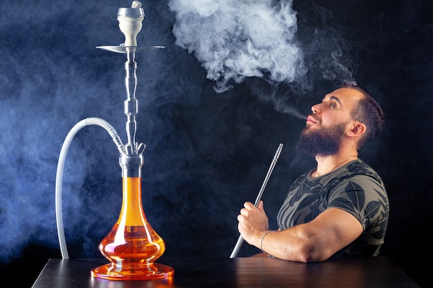 Bearded young man smoking shisha in a dark night club close up