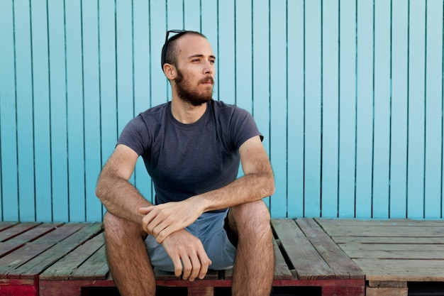 Bearded young man wearing sunglasses over the head is confidently looking away at the Isla Canela beach with a blue wooden wall background