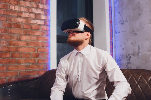 Bearded young man wearing virtual reality goggles in modern coworking studio. 