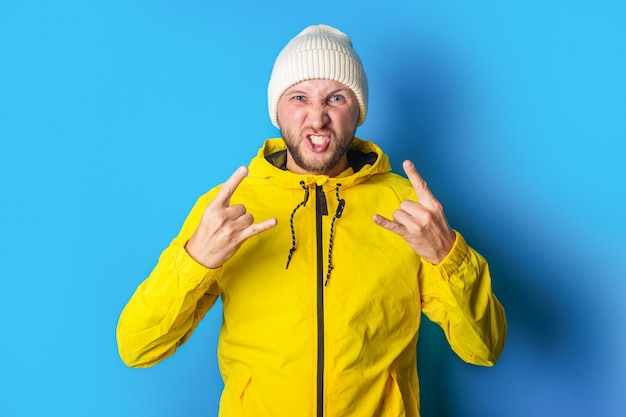 A bearded young man in a yellow jacket shows a rocker goat, a horn on a blue background.