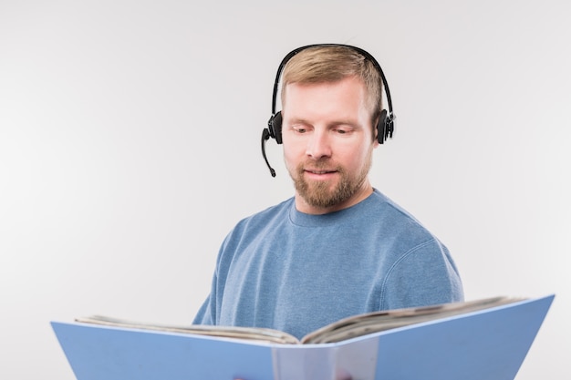 Bearded young operator with headset consulting clients while looking through papers in folder in isolation