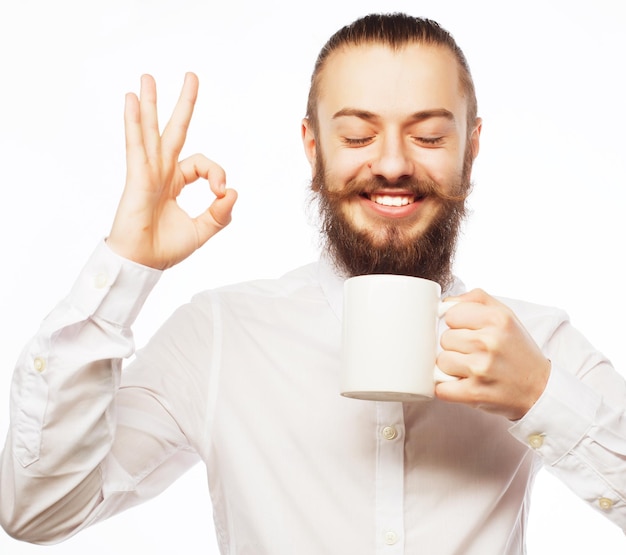 Beardige jonge man drinkt koffie en toont duim omhoog.