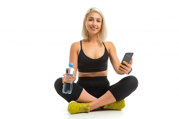 Красивая спортивная кавказская девушка сидит на полу, скрестив ноги, держит мобильный телефон и бутылку воды, общается с друзьями в изоляции