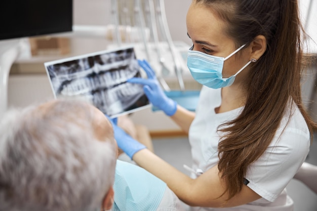 Красивая брюнетка женщина-дантист в маске держит рентгеновский снимок челюстей, показывая его своему пациенту