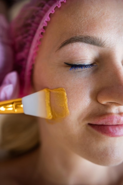 A beautician applies a golden mask with a brush on a young beautiful face of a female client in a beauty clinic Spa salon Cosmetologist doing facial skin care procedure