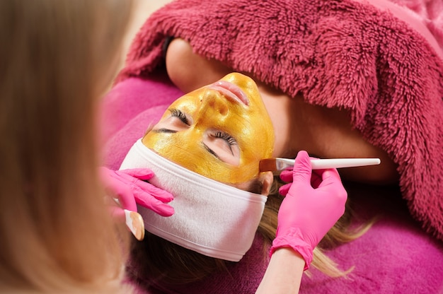 美容師は女性の顔に黄金のマスクを適用します
