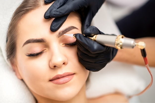 Photo beautician applying permanent makeup on eyebrows by tattoo machine tool