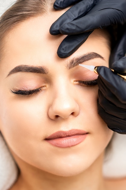 Beautician applying permanent makeup on eyebrows of young woman by special tattoo machine tool