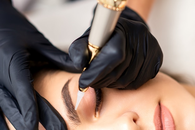 Beautician applying permanent makeup on eyebrows of young woman by special tattoo machine tool