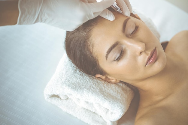 Beautician doing beauty procedure with syringe to face of young brunette woman in sunny clinic. Cosmetic medicine and surgery, beauty injections.