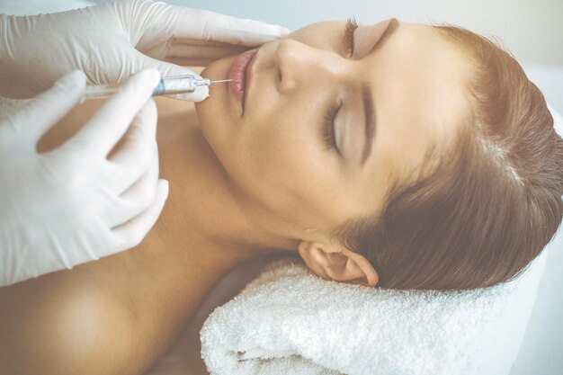 Beautician doing beauty procedure with syringe to face of young\
brunette woman in sunny clinic. cosmetic medicine and surgery,\
beauty injections