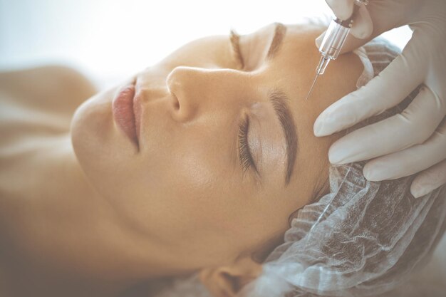 Beautician doing beauty procedure with syringe to face of young\
brunette woman in sunny clinic. cosmetic medicine and surgery,\
beauty injections