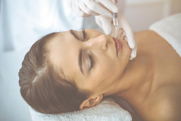 Beautician doing beauty procedure with syringe to face of young brunette woman in sunny clinic. Cosmetic medicine and surgery, beauty injections.