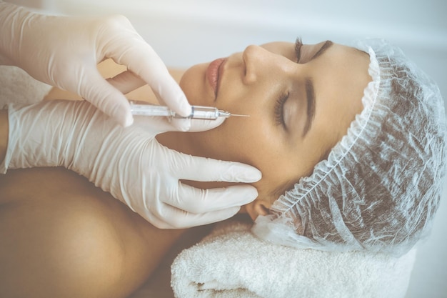 beautician doing beauty procedure with syringe to face of young brunette woman in sunny clinic. cosmetic medicine and surgery, beauty injections.