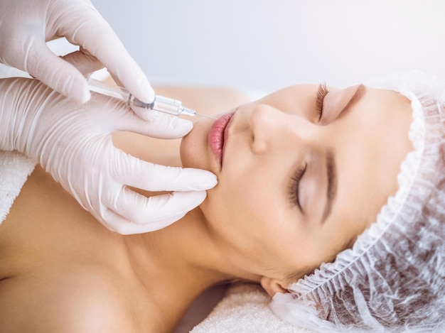 Beautician doing beauty procedure with syringe to face of young brunette woman in sunny spa center. Cosmetic medicine and surgery, beauty injections.