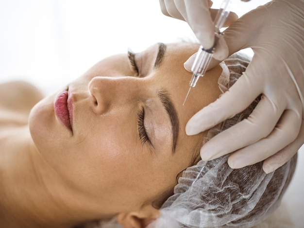 Beautician doing beauty procedure with syringe to face of young brunette woman in sunny spa center. Cosmetic medicine and surgery, beauty injections.
