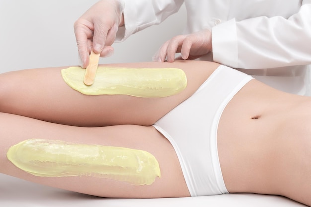 Beautician in gloves applying green hot wax on slim woman legs using spatula while woman lying