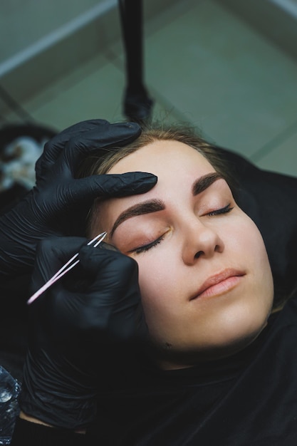 beautician hand doing brow permanent makeup on an attractive female face