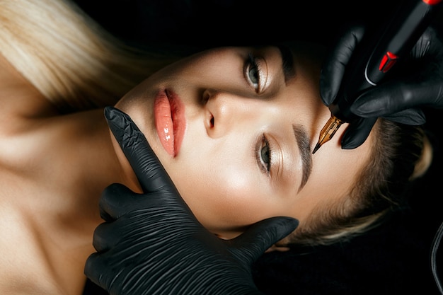 Рука косметолога делает татуировку бровей на женских бровях