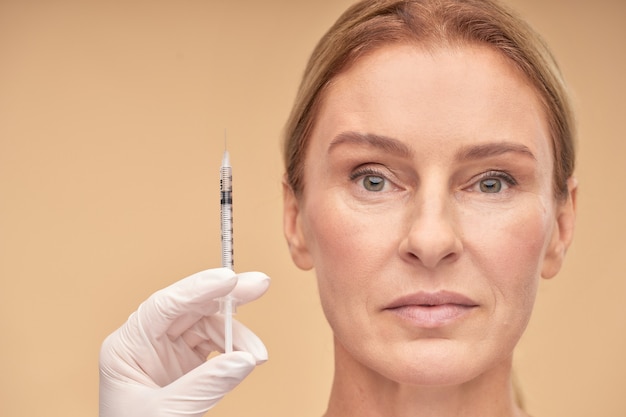 Photo beautician hand in white glove holding syringe near mature woman face over beige background