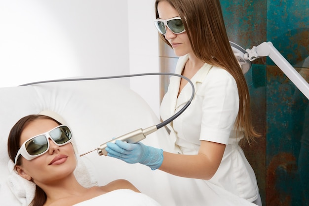 Beautician holding laser for rejuvenation, vessels or hair removal near beautiful woman face in protection glasses at spa salon