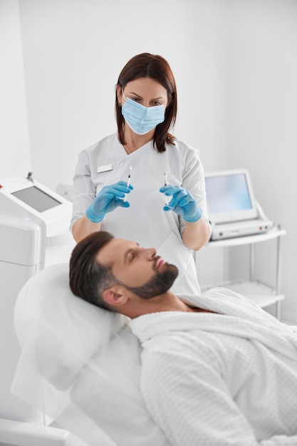 Косметолог держит шприцы с наполнителем для подтяжки кожи рядом с мужчиной средних лет в клинике