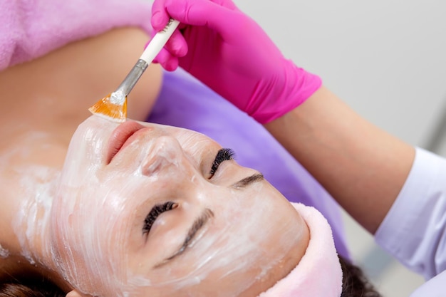 Beautician makes a face clay mask against acne on the face of a woman to rejuvenate the skin Cosmetology treatment of problem skin on the face and body