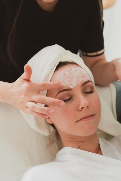 Beautician makes facial and massage and cosmetic cream to adorable young woman with a towel on her head