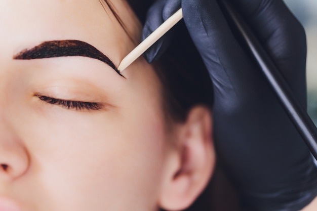 beautician- makeup artist applies paint henna on previously plucked, design, trimmed eyebrows in a beauty salon in the session correction. professional care for face.