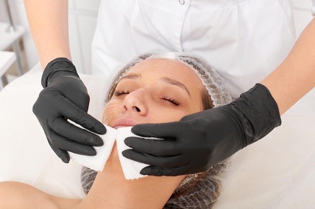 Beautician removing facial cream mask from woman face skin with cosmetic wipes in beauty salon