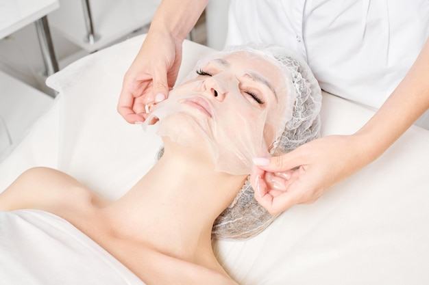 Beautician smoothes sheet mask on woman face for rehydrate face skin procedure in beauty salon