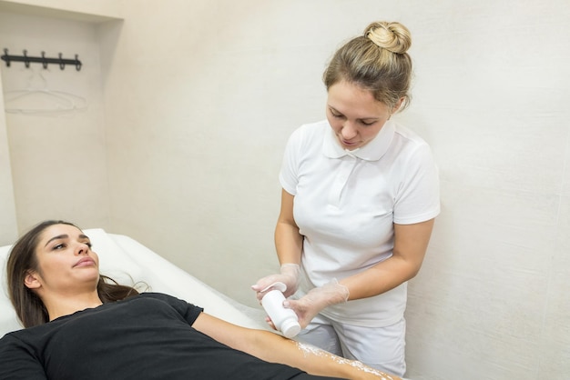 The beautician sprinkles and distributes talcum powder on the arm of a young girl before the epilation procedure The girl lies on the couch in the beauty salon doing the sugaring procedure