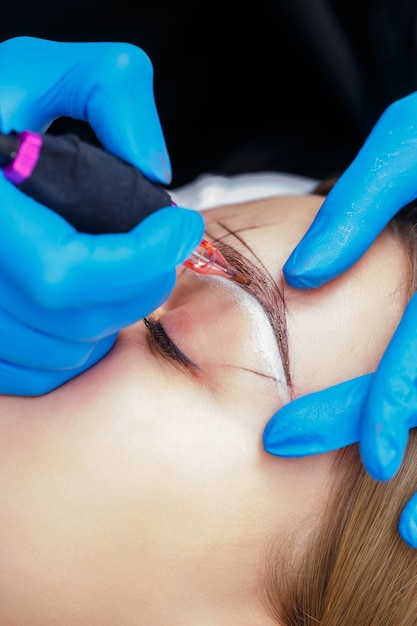 Photo beautician tattooing a woman's eyebrows using special equipment during permanent make-up, close-up.