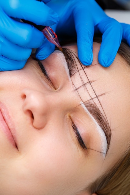 Beautician tattooing a woman's eyebrows using special equipment during permanent make-up, close-up.