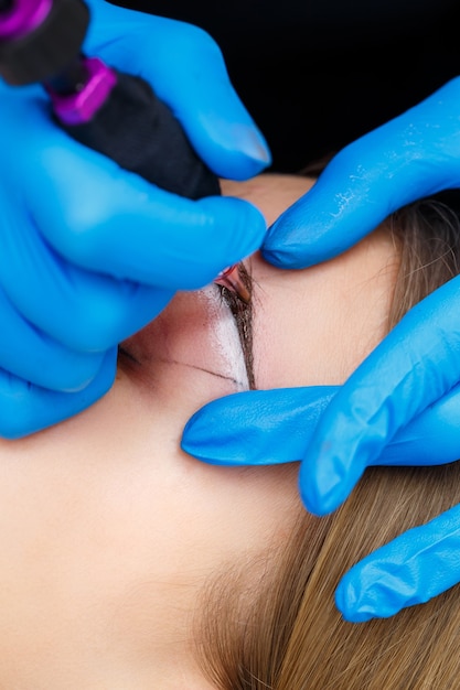 Косметолог татуирует женские брови с помощью специального оборудования во время перманентного макияжа, крупным планом.
