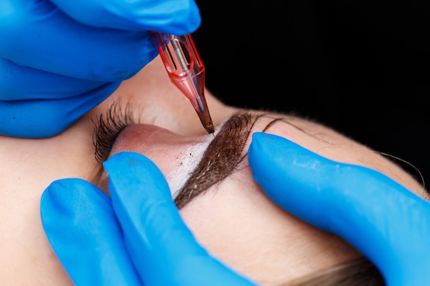 Beautician tattooing a woman's eyebrows using special equipment during permanent make-up, close-up.