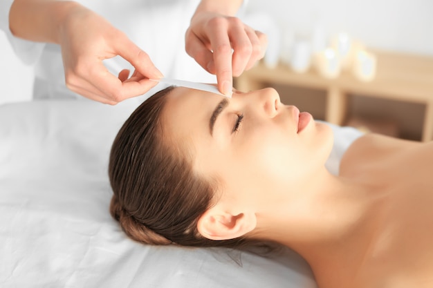 Beautician waxing young woman's eyebrows in spa center