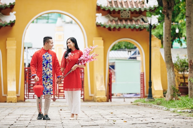 Beautifujl young just married couple in traditional clothes holding hands and walking on the street with bunch of peach branches