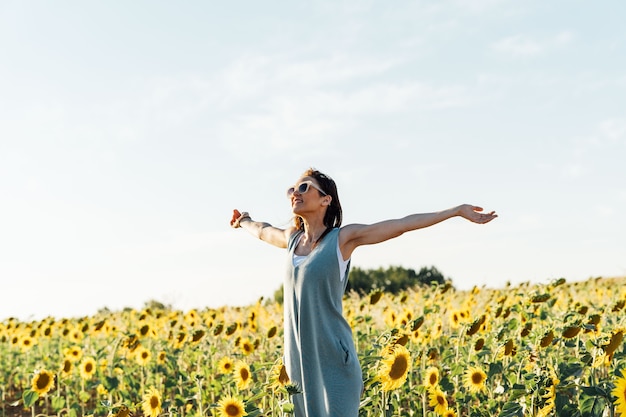 Beautiful 40 year old woman breathing fresh air in sunflower field with open arms and sunglasses