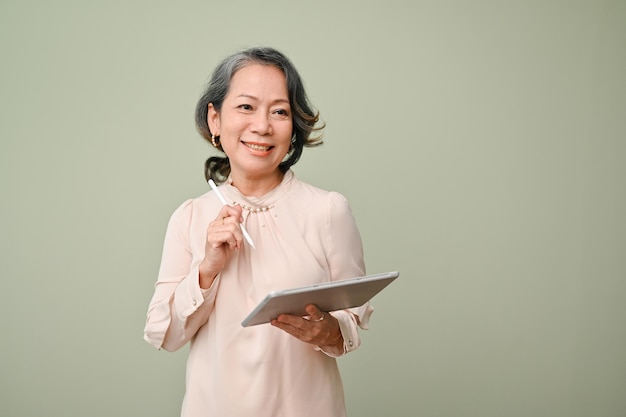 Beautiful 60s agedasian woman holding a tablet and pen standing isolated over green background