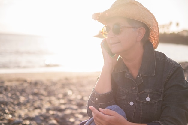 美しい大人の白人女性が海のあるビーチで電話で話します。夏の接続コンセプトの日没時間。自然とテクノロジーをお楽しみください