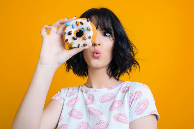 Photo beautiful adult female dressed in a shirt with donuts looking astonished through a donut in front of a yellow studio wall.