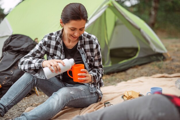 Bella ragazza adulta che beve tè in campeggio nella foresta con tenda sullo sfondo e giovane donna sorridente...